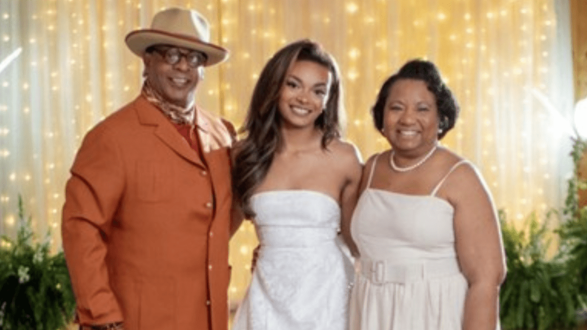A smiling man in a rust-colored suit and cream hat stands beside a young woman in a strapless white gown and an older woman in a light beige dress, posing together in front of a warm backdrop of golden string lights and greenery, suggesting a bridal or wedding setting.